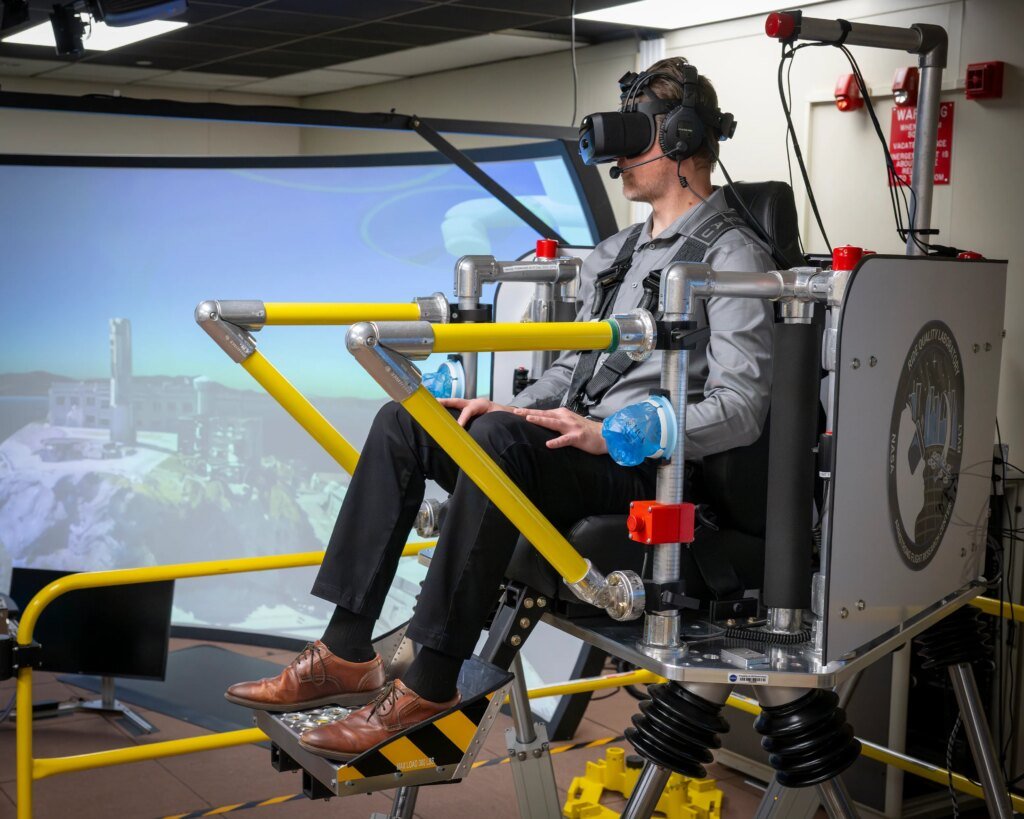 A brown and yellow platform holding a seat that is raised up in the air is holding one passenger. The passenger is wearing virtual reality goggles and a harness. There is a TV monitor to the right of the monitor showing the words “liftoff” and a second screen behind the passenger.
